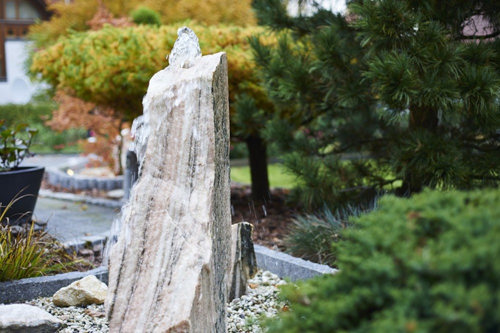 Wasserspiel mit Naturstein in der Gartengestaltung Natürliches Wasserspiel aus aufrechtem Naturstein zwischen Kies, Gehölzen und Gartenpflanzen